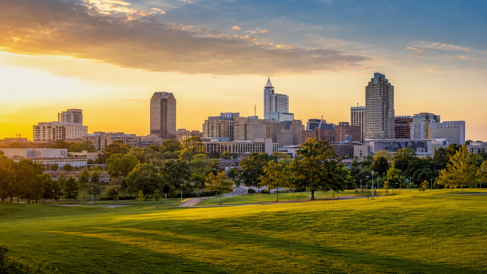 Raleigh skyline at golden hour with downtown buildings, green park space, open lawn and sunset sky in North Carolina.