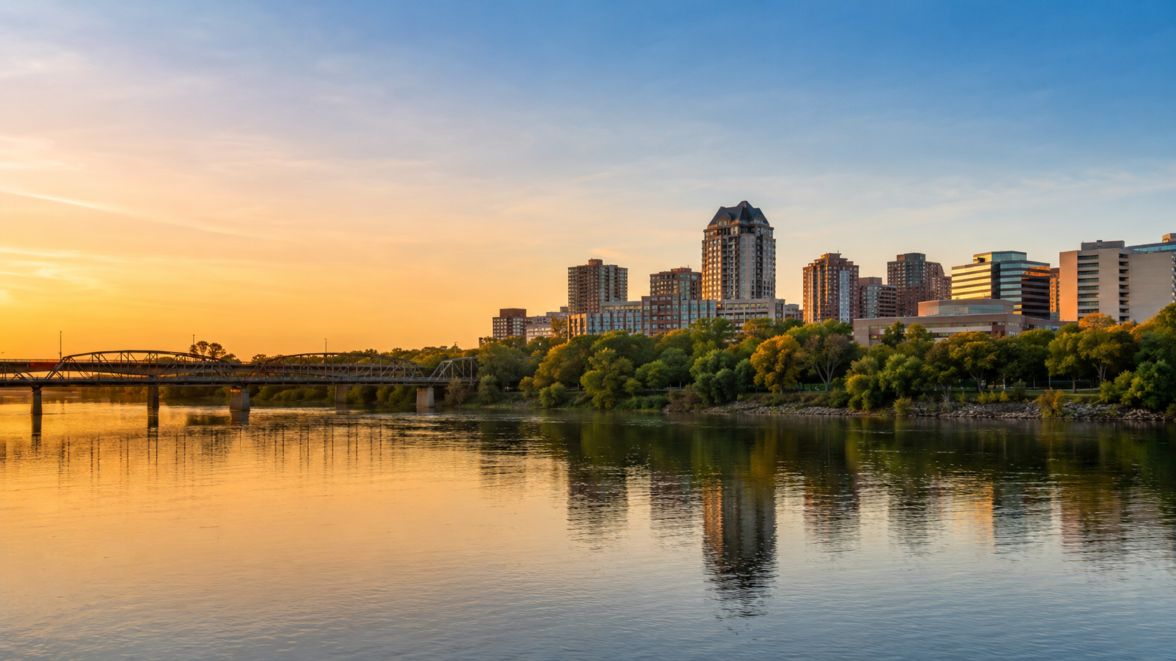 Saskatoon skyline at golden hour with South Saskatchewan River, downtown buildings, bridge and waterfront reflections.