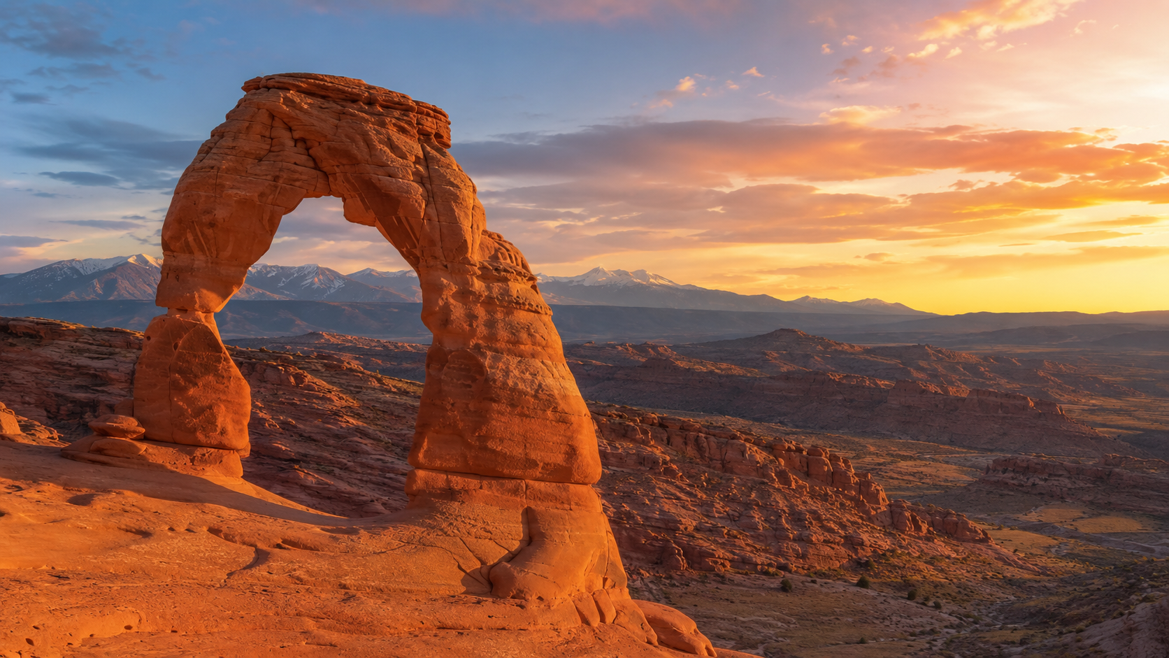 Utah desert landscape at golden hour with Delicate Arch, red sandstone canyons, distant mountains and sunset sky.